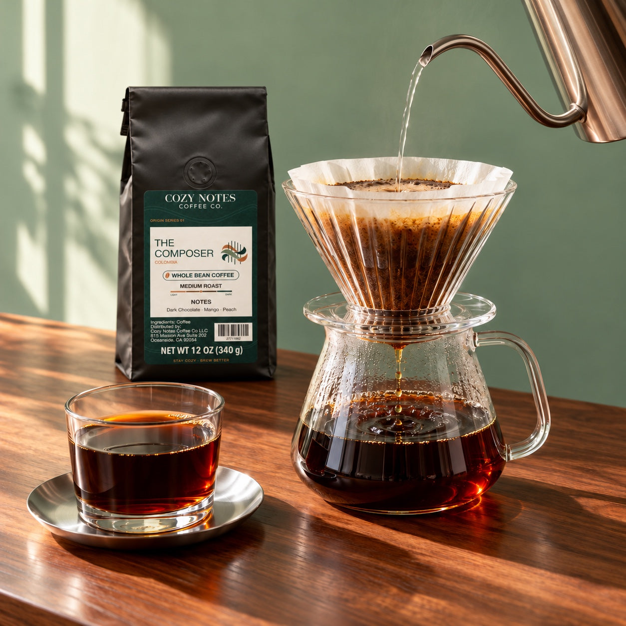 Fresh pour-over coffee in glass, with The Composer coffee bag on a sunlit walnut counter.