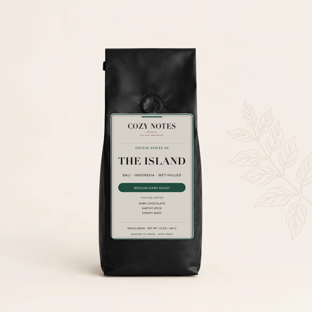 A black bag of Cozy Notes 'The Island' medium dark roast coffee sits on a warm off-white background with a subtle botanical line illustration on the right.