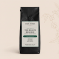 A black coffee bag from Cozy Notes, labeled 'The Bloom Reserve', standing upright against a light beige background with a subtle botanical line drawing on the right.
