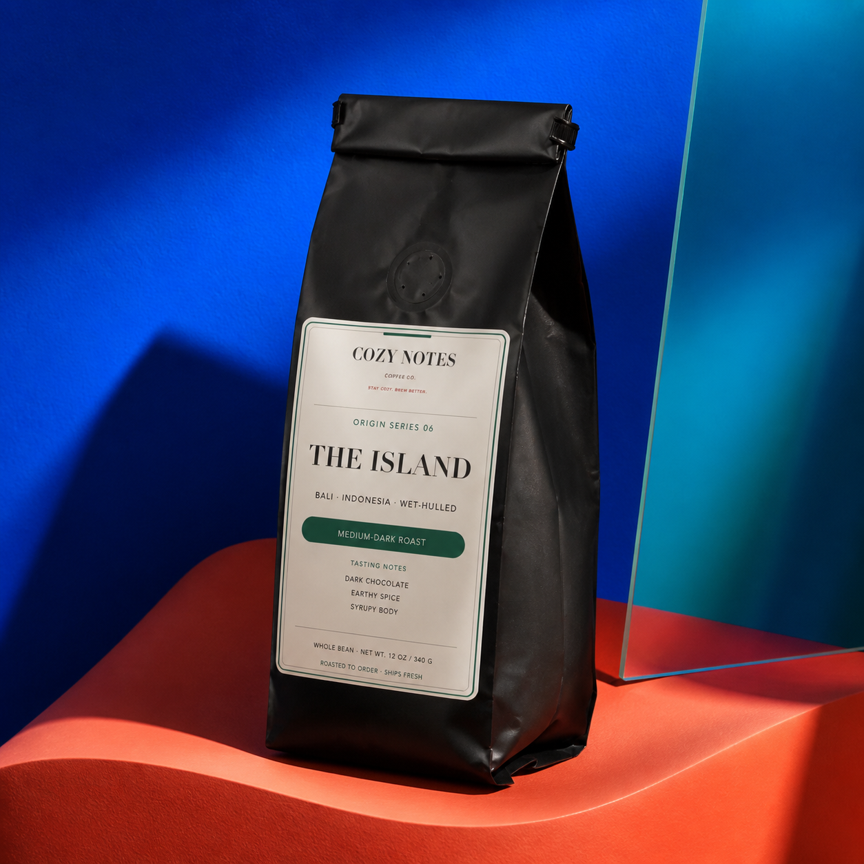 A black coffee bag labeled Cozy Notes The Island Medium Dark Roast stands on a bright orange platform against a vivid blue background with a translucent blue panel.