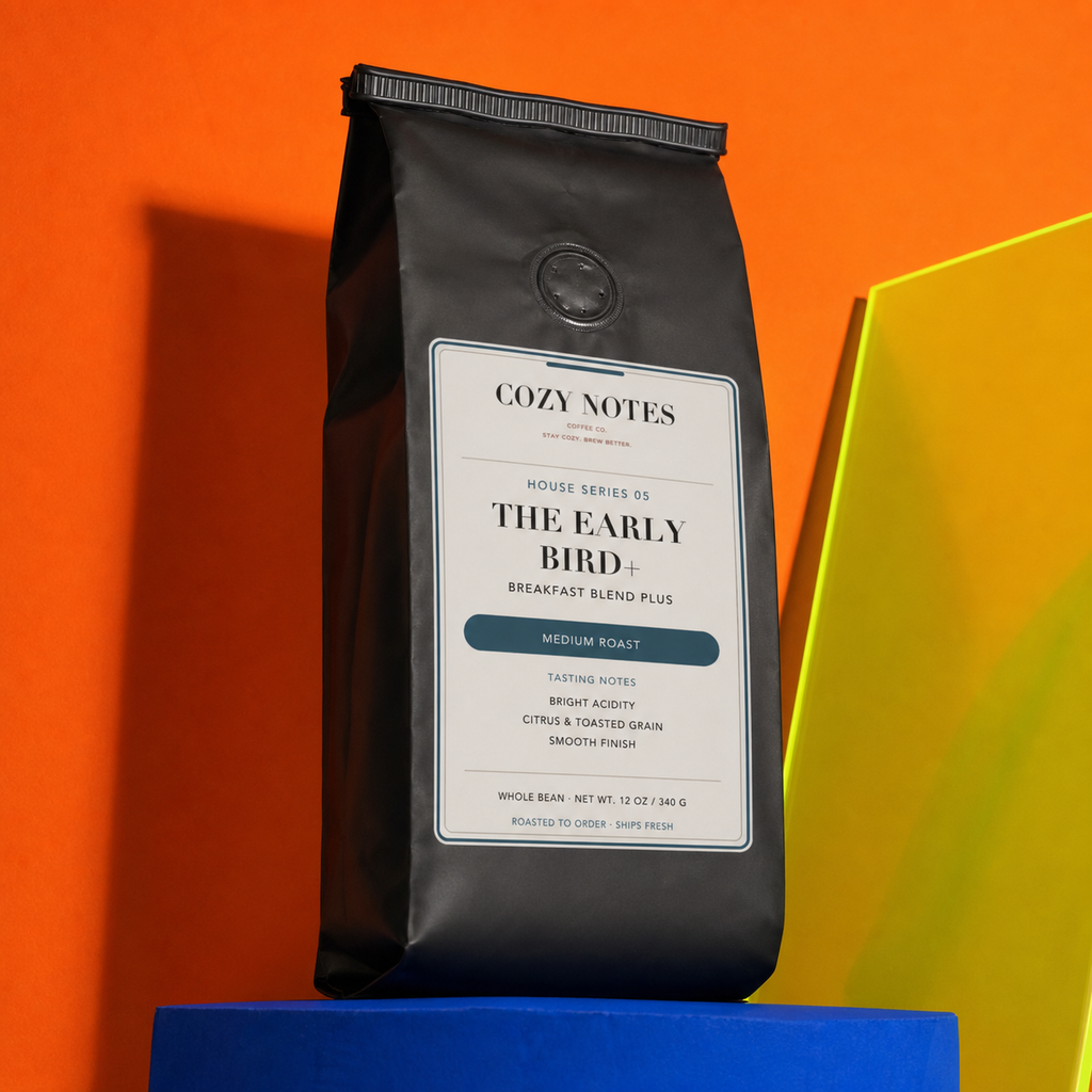 A black bag of Cozy Notes Coffee Roasters 'The Early Bird+' medium roast coffee stands upright on a blue pedestal against a bright orange background, with a yellow geometric shape partially visible on the right.
