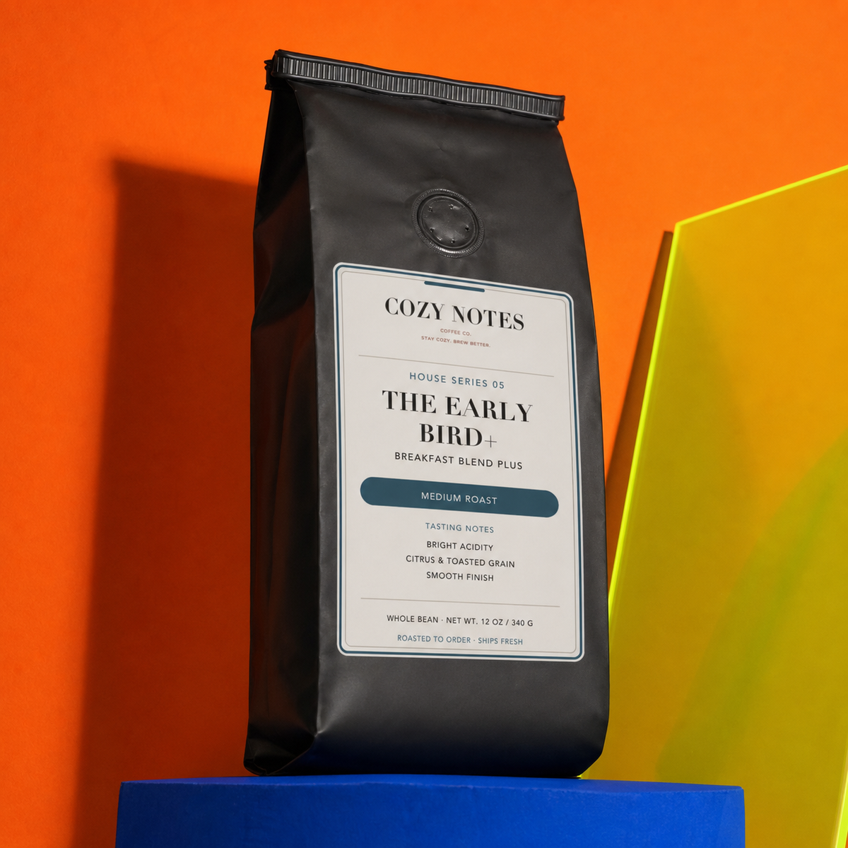 A black bag of Cozy Notes Coffee Roasters 'The Early Bird+' medium roast coffee stands upright on a blue pedestal against a bright orange background, with a yellow geometric shape partially visible on the right.