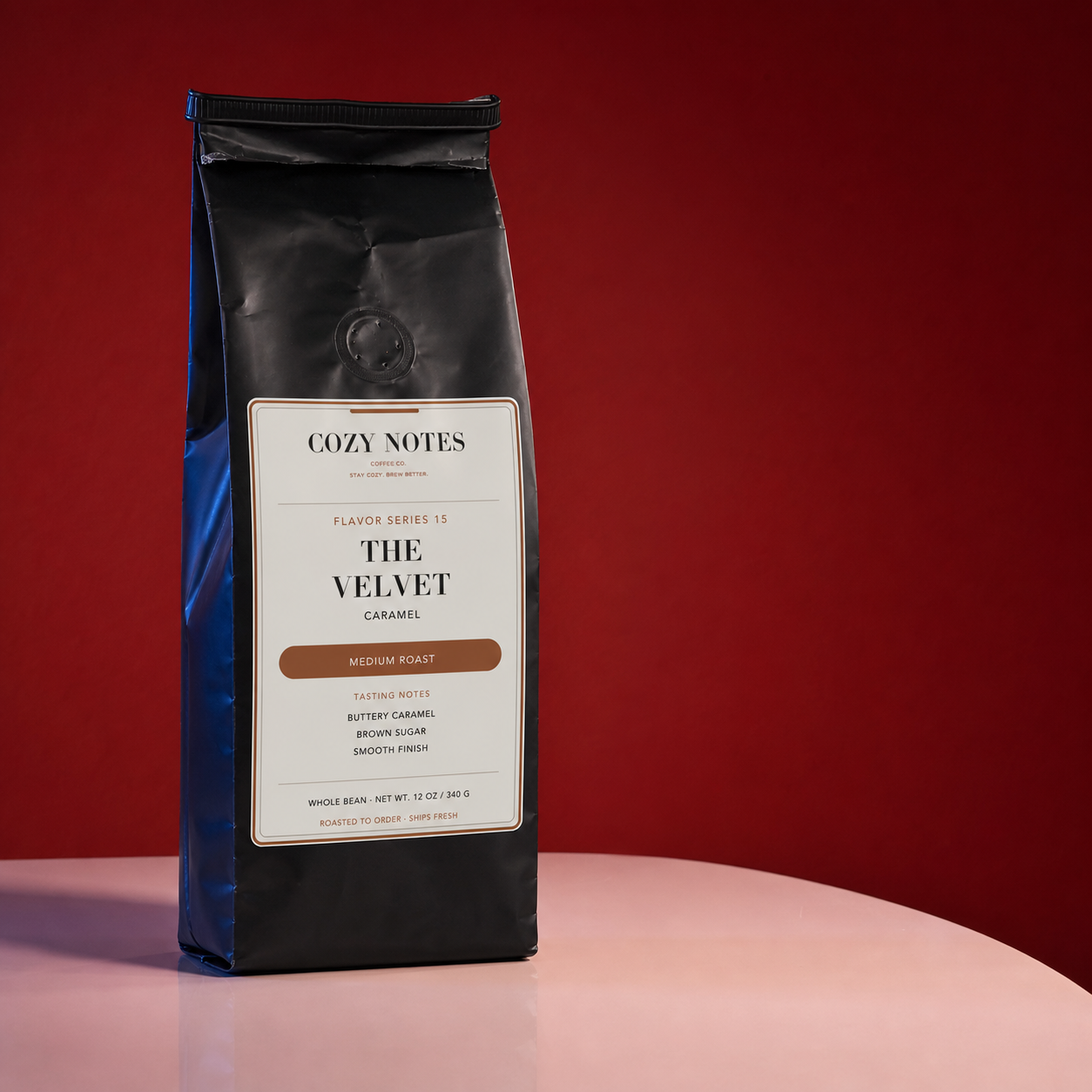 A bag of Cozy Notes Coffee Roasted Coffee, specifically 'The Velvet Caramel' medium roast, standing on a light pink surface against a dark red background.