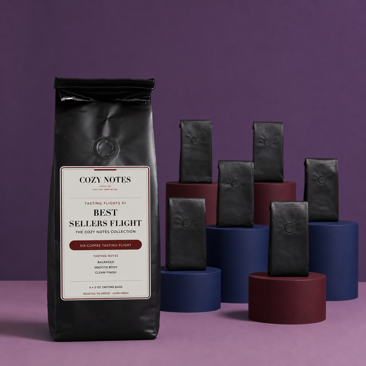 A black bag of Cozy Notes coffee stands prominently in the foreground on a purple studio background, with several smaller black product bags displayed on blue and burgundy cylindrical pedestals behind it.
