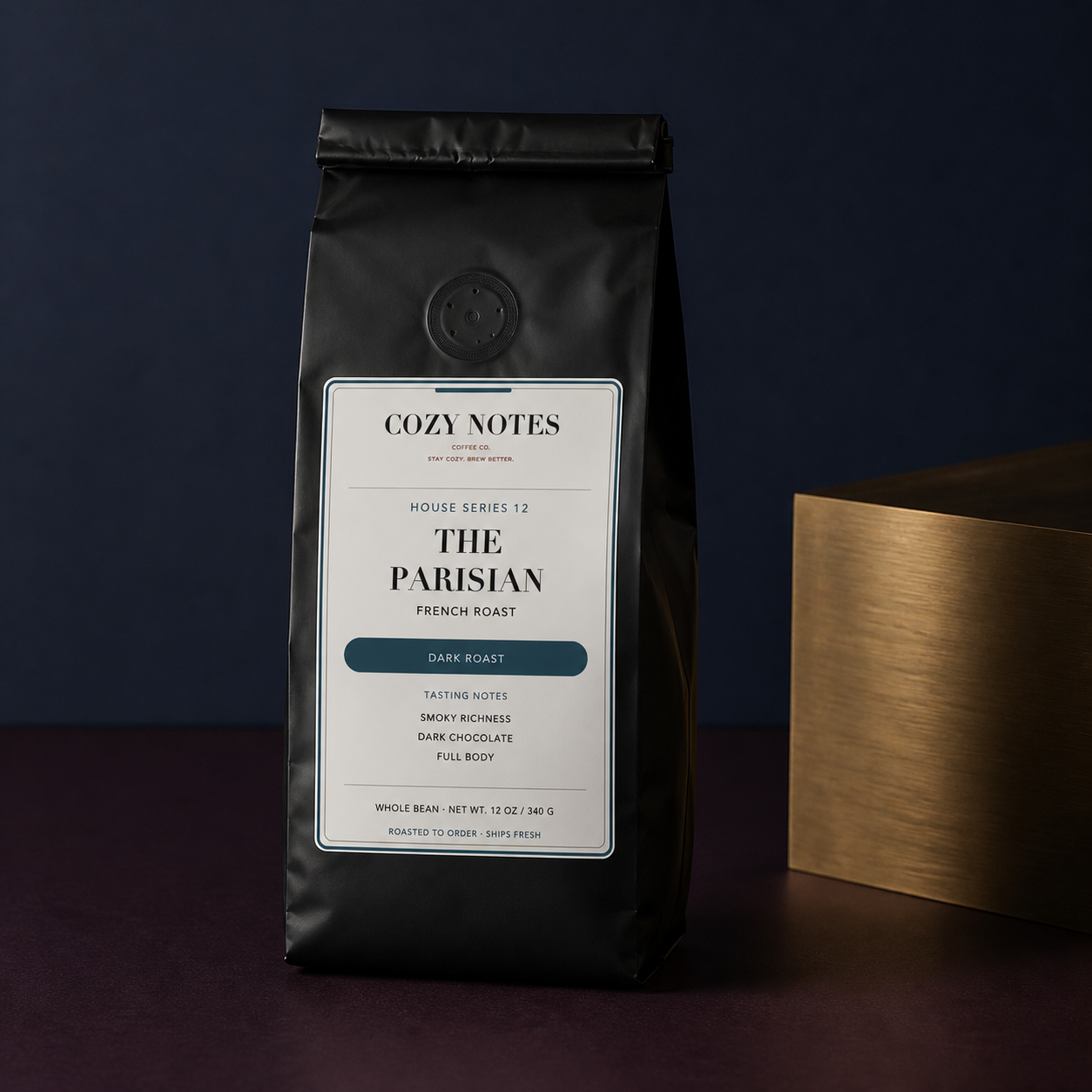 A black bag of Cozy Notes The Parisian Dark Roast coffee stands on a dark surface against a deep navy background, with a gold box partially visible on the right.