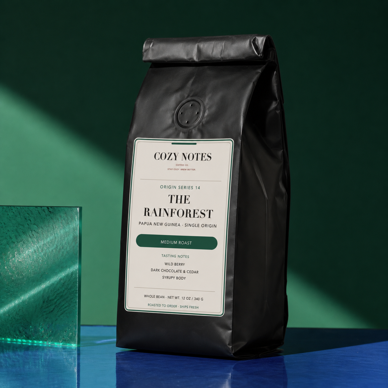 A matte black coffee bag labeled Cozy Notes The Rainforest stands upright on a blue reflective surface against a dark green background. A green glass block is visible at the left edge.