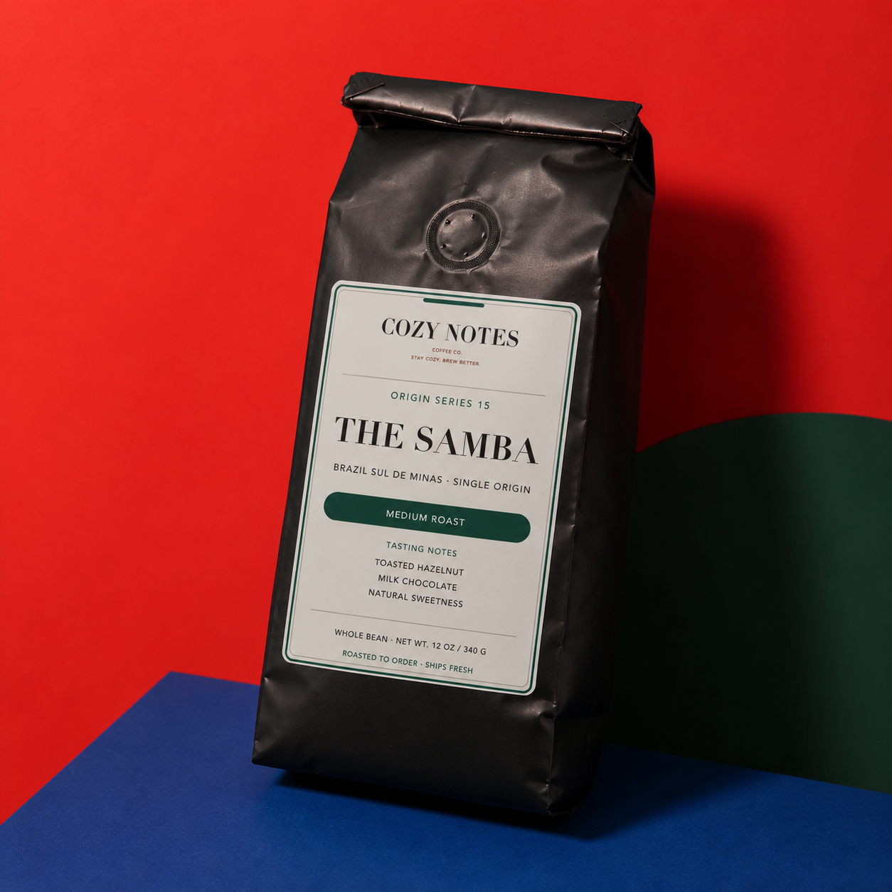 A black coffee bag labeled 'Cozy Notes The Samba' stands upright on a colorful studio setup with a bright red background and blue and green surfaces at the bottom.