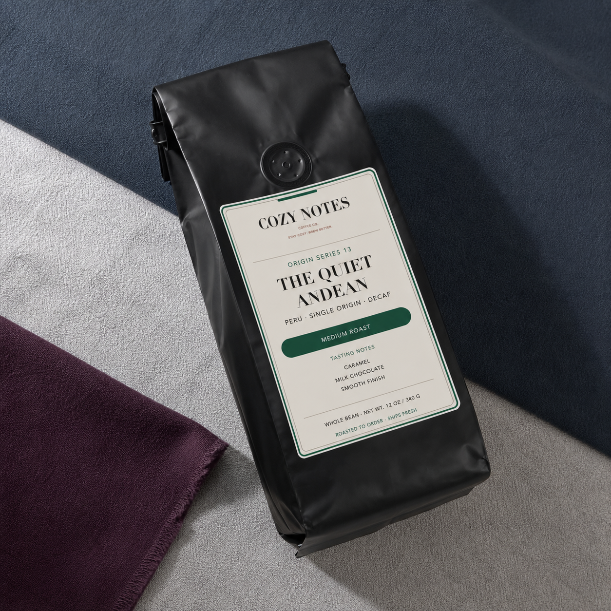 A black coffee bag labeled Cozy Notes The Quiet Andean lies on a gray surface with folded fabric in the background and foreground.